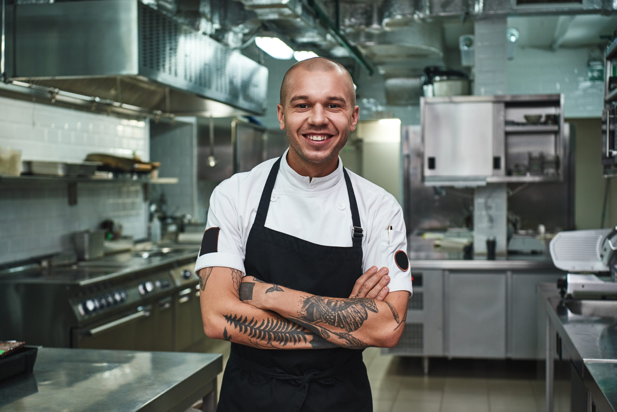 Cheerful young chef in apron keeping tattooed arms crossed and smiling while standing in a restaurant kitchen